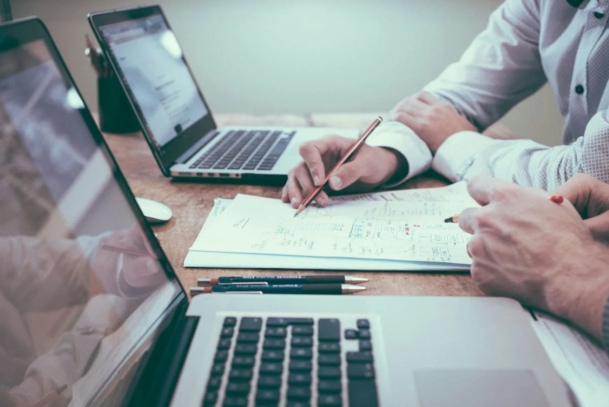 Two people reviewing documents at a table with laptops and pens.