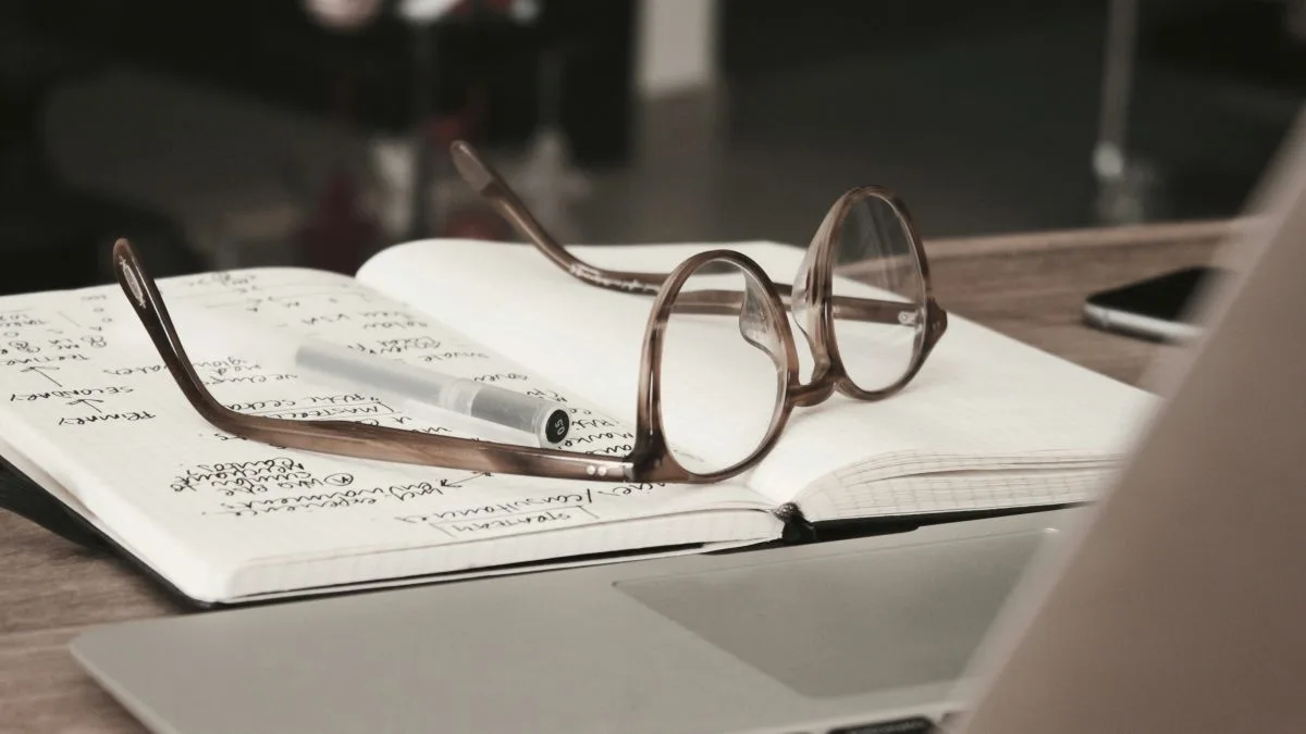 Glasses and pen on an open notebook next to a laptop.