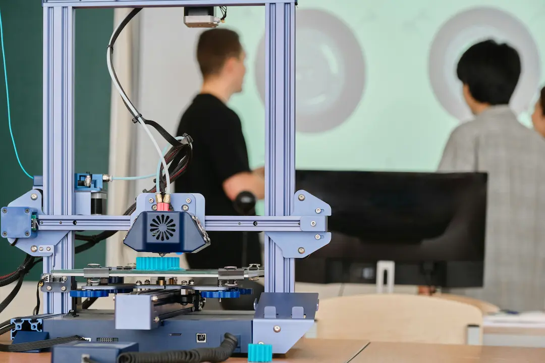 Open 3D printer printing an object, blurred people in the background.