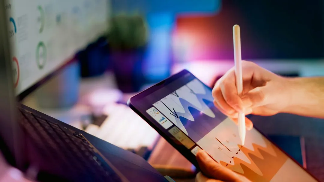 Hand using stylus on tablet displaying charts next to a laptop.