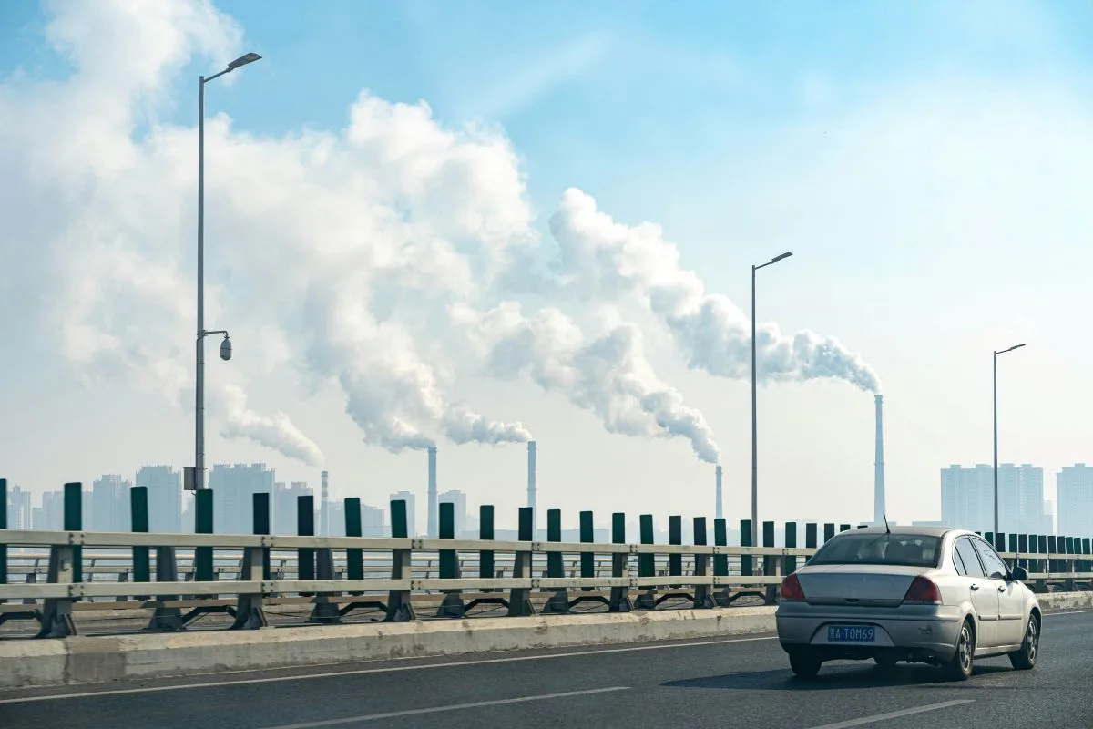 Car driving on highway with factory smokestacks emitting smoke in the background.